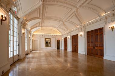 Open hardwood space with large mirror at the end of the hallways, large windows on the left side, and three large wood doors on the right. 