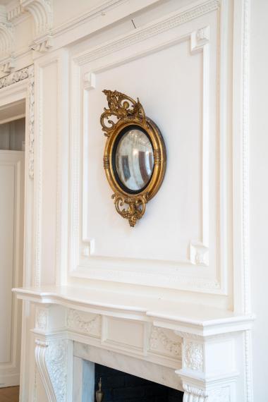 Gold circular decorative mirror hangs over a mantle with ornate moulding