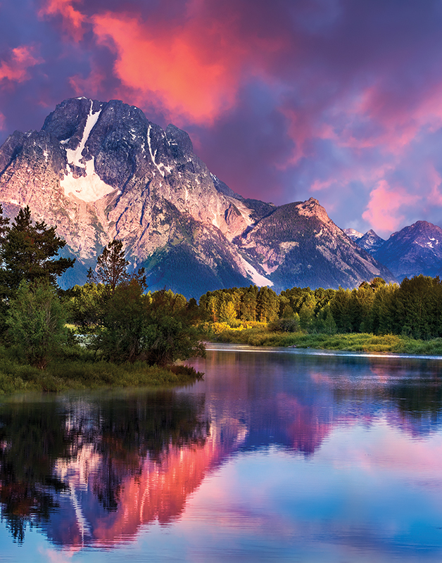 Teton mountain range at sunset with brilliant blues, pinks, and oranges with a lake in front of it and green lush trees.