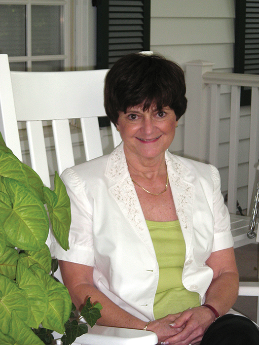 Brunette woman with green shirt and white blazer sitting on a white rocking chair on a porch. 