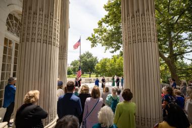 Women at a podium and members surround as the United States Flag is raised on the flag pole. 