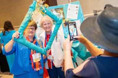 Women poses at a photo booth with a turquoise frame over their faces while another woman takes a photo on her phone.