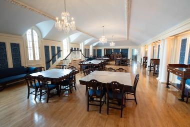 Large wooden floor room and blue fabric panelled walls that has five wooden tables with chairs at them while on the ceiling three glass chandeliers 
