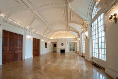 Open hardwood floors with mantle, fire place, and circle mirror at the end of the hall, large windows on the right side, and two large sliding wood doors on the left. 
