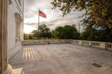 unset casting long shadows on a paved area near a building with an american flag waving in the background.