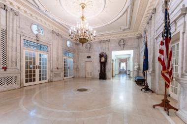 marble room with busts on the wall, a grandfather clock at the end of the room, and a chandelier hanging. 