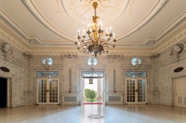 A marble room with an open door and a gold chandelier hanging. 