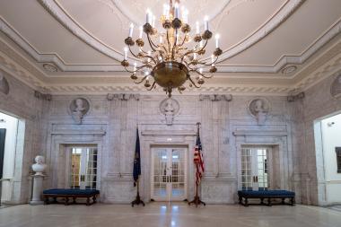 A marble room with a door at the end of the hallway and a gold chandelier hanging.