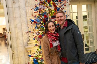 A man and a woman smiling in front of a patriotic Christmas tree. 